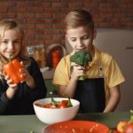 Cute girl and boy with fresh pepper and broccoli in hands standing near desk with assorted vegetables and salad bowl during vegetarian meal preparation