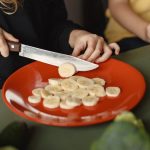 Two children preparing sliced bananas on a red plate in a kitchen setting.